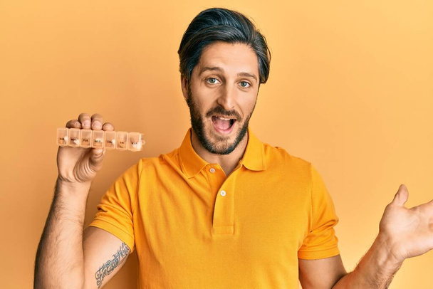 Young hispanic man holding pill organizer celebrating achievement with happy smile and winner expression with raised hand  - 写真・画像