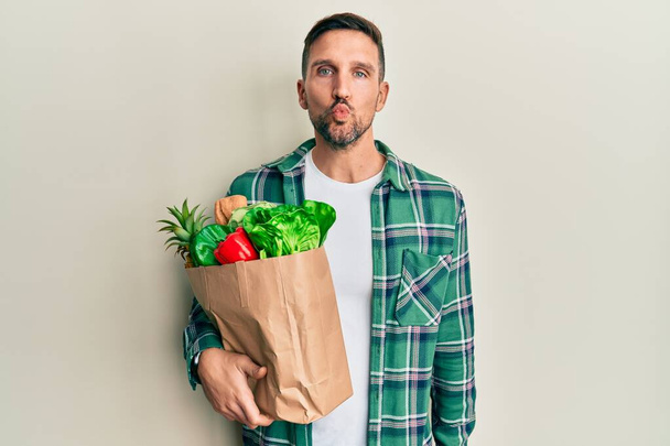 Handsome man with beard holding paper bag with groceries looking at the camera blowing a kiss on air being lovely and sexy. love expression.  - Фото, изображение