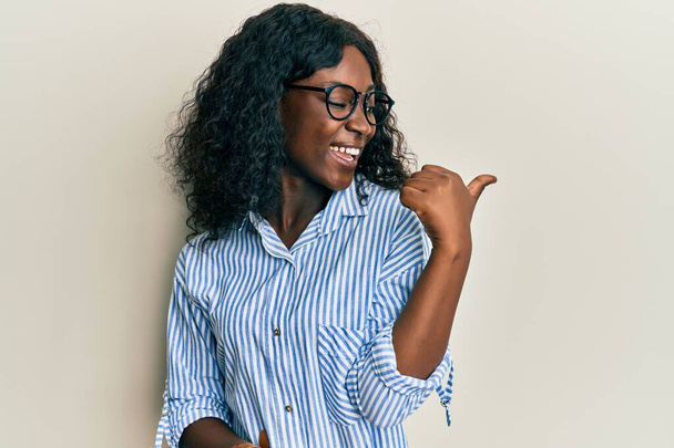 Beautiful african young woman wearing casual clothes and glasses pointing thumb up to the side smiling happy with open mouth  - Φωτογραφία, εικόνα