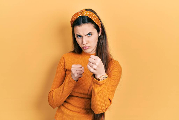 Giovane adolescente bruna con indosso maglione giallo casual pronto a combattere con gesto di difesa pugno, viso arrabbiato e sconvolto, paura del problema  - Foto, immagini