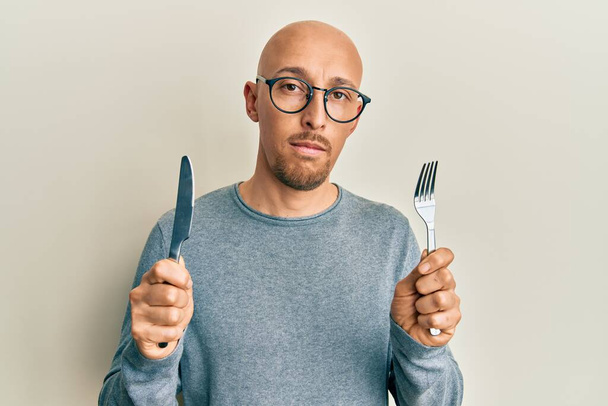 Homme chauve avec barbe tenant fourchette et couteau prêt à manger détendu avec une expression sérieuse sur le visage. simple et naturel regardant la caméra.  - Photo, image