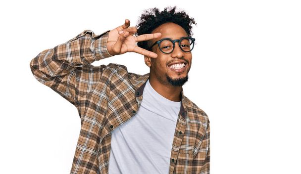 Young african american man with beard wearing casual clothes and glasses doing peace symbol with fingers over face, smiling cheerful showing victory  - Φωτογραφία, εικόνα