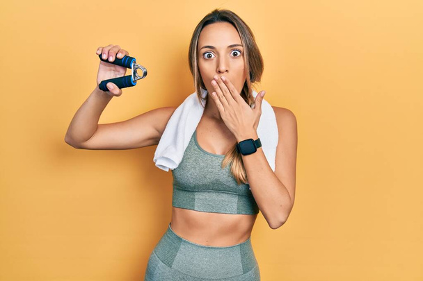 Beautiful hispanic woman training muscle with hand grip covering mouth with hand, shocked and afraid for mistake. surprised expression  - Photo, Image