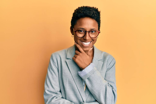 Jong Afrikaans Amerikaans meisje draagt een zakenjasje en een bril die zelfverzekerd kijkt naar de camera die glimlacht met gekruiste armen en hand geheven op kin. positief denken.  - Foto, afbeelding