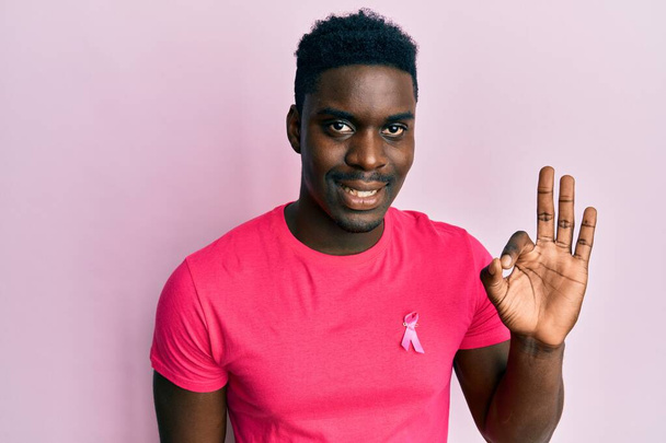 Hombre negro guapo con cinta de cáncer rosa unido a la camisa haciendo signo de ok con los dedos, sonriendo gesto amistoso excelente símbolo  - Foto, Imagen