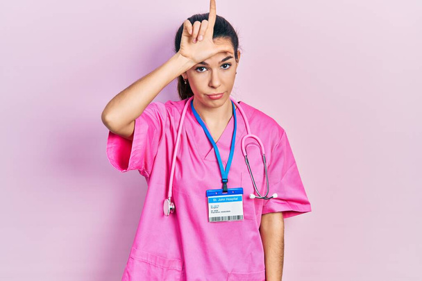 Jeune femme brune portant l'uniforme de médecin et stéthoscope se moquant des gens avec les doigts sur le front faisant geste de perdant se moquant et insultant.  - Photo, image