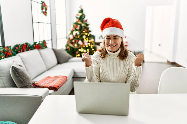 Keski-ikäinen nainen yllään joulupukki hattu kannettavan tietokoneen avulla huutaa ylpeä, juhlii voittoa ja menestystä hyvin innoissaan nosti kädet  - Valokuva, kuva