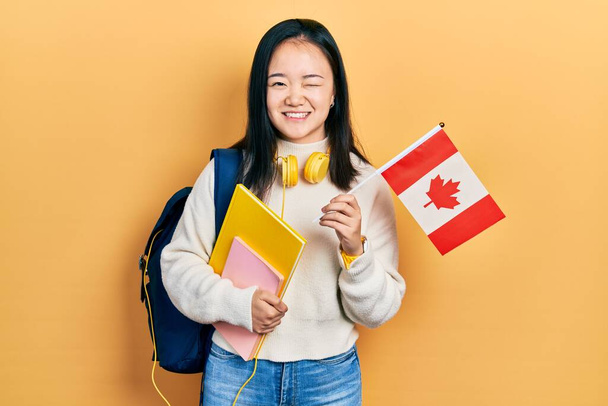 Joven estudiante de intercambio de chicas chinas sosteniendo la bandera de Canadá guiño mirando a la cámara con expresión sexy, cara alegre y feliz.  - Foto, imagen