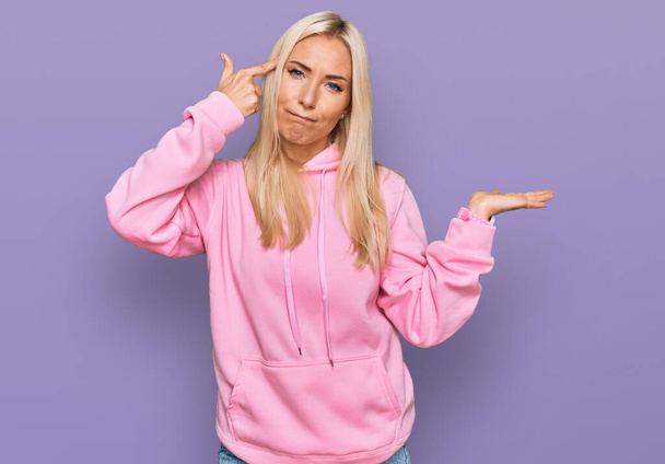 Young blonde woman wearing casual sweatshirt confused and annoyed with open palm showing copy space and pointing finger to forehead. think about it.  - Photo, Image
