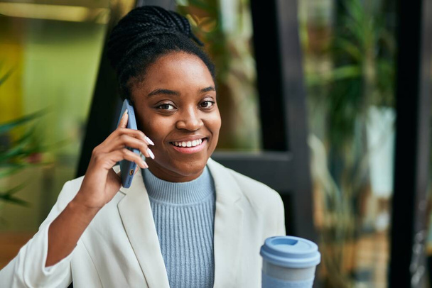 Young african american businesswoman talking on the smartphone and drinking coffee at the city. - 写真・画像