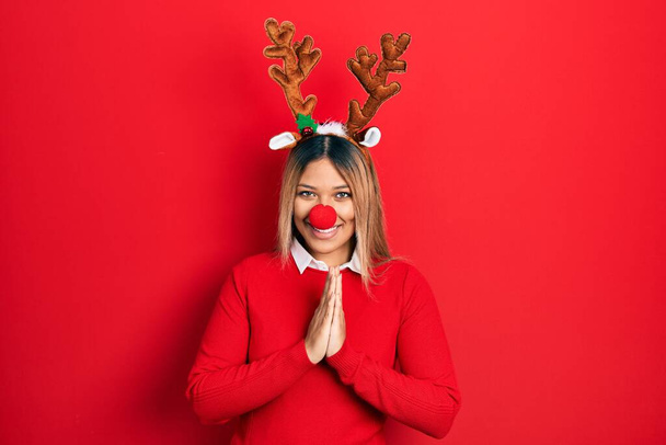 Красивая латиноамериканка в рождественской шляпе оленя и с красным носом, молящая руки вместе, просит прощения, уверенно улыбаясь..  - Фото, изображение