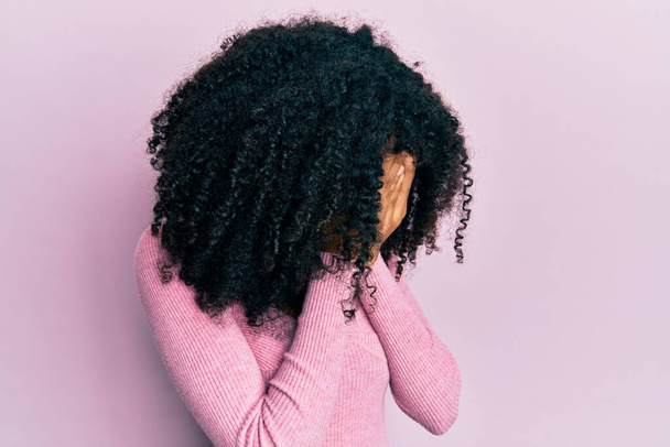 Femme afro-américaine aux cheveux afro portant une chemise rose décontractée avec une expression triste couvrant le visage avec les mains tout en pleurant. concept de dépression.  - Photo, image