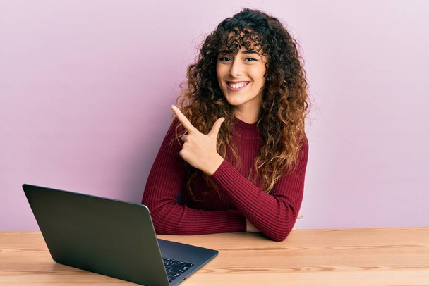 Junges hispanisches Mädchen, das mit Computer-Laptop arbeitet, lächelt fröhlich und zeigt mit der Hand und dem Finger zur Seite  - Foto, Bild