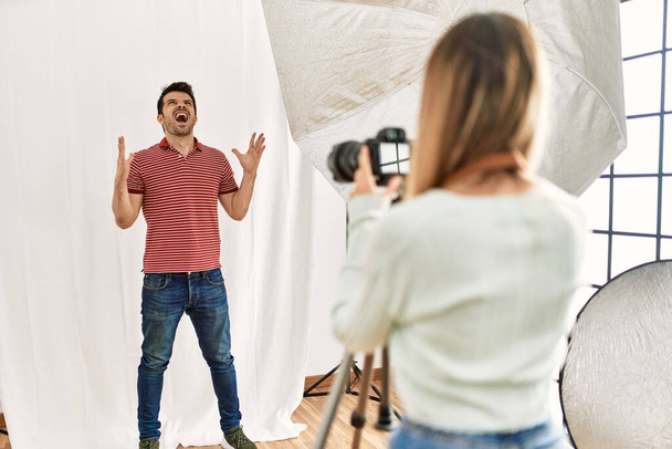 Kobieta fotograf mówi zdjęcia mężczyzny udającego modelkę w studiu fotograficznym szalony i szalony krzyczy i krzyczy z agresywną ekspresją i ramionami podniesione. koncepcja frustracji.  - Zdjęcie, obraz