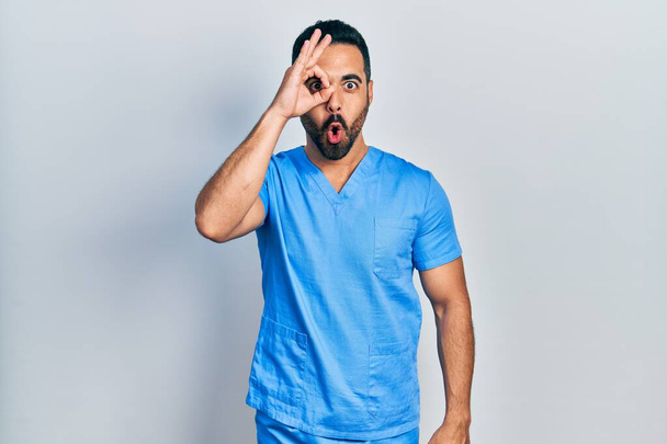 Przystojny Latynos z brodą w niebieskim męskim mundurze pielęgniarki wykonujący ok gestem wstrząśnięty zaskoczoną twarzą, okiem patrzącym przez palce. niewierząca ekspresja.  - Zdjęcie, obraz