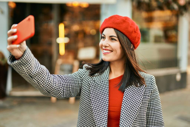 Młoda Latynoska uśmiecha się szczęśliwa robiąc selfie przez smartfona w mieście. - Zdjęcie, obraz