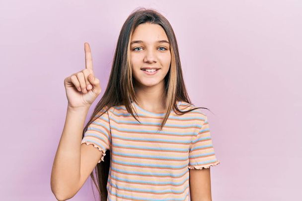 Νεαρό μελαχρινό κορίτσι που φοράει casual ριγέ μπλουζάκι χαμογελώντας με μια ιδέα ή ερώτηση δείχνοντας το δάχτυλο προς τα πάνω με χαρούμενο πρόσωπο, νούμερο ένα  - Φωτογραφία, εικόνα