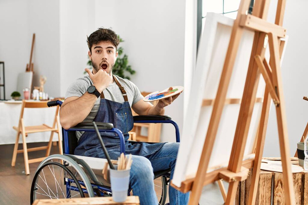 Junger hispanischer Mann, der im Rollstuhl im Kunstatelier sitzt, überrascht mit dem Finger zur Seite zeigend, offener Mund erstaunter Ausdruck.  - Foto, Bild