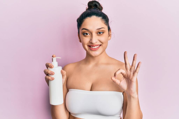 Belle femme du Moyen-Orient tenant crème hydratante cosmétique visage faisant signe ok avec les doigts, sourire geste amical excellent symbole  - Photo, image