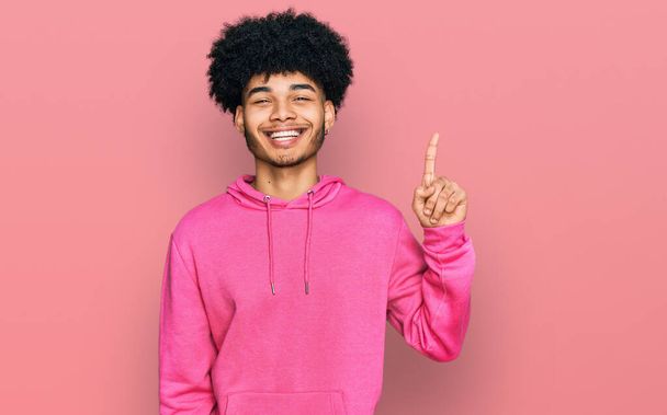若いですアフリカ系アメリカ人男性とともにアフロ髪身に着けていますカジュアルなピンクのスウェットシャツ表示され、指の番号1で指摘しながら、自信と幸せ.  - 写真・画像