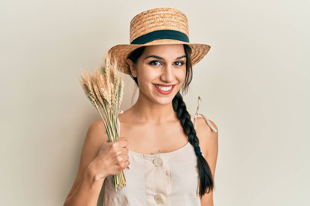 Młoda Latynoska kobieta w letniej czapce z kolczastą pszenicą wyglądająca pozytywnie i szczęśliwie stojąca i uśmiechnięta z ufnym uśmiechem pokazującym zęby  - Zdjęcie, obraz