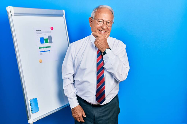 Senior man with grey hair standing by business blackboard looking confident at the camera smiling with crossed arms and hand raised on chin. thinking positive.  - Photo, Image