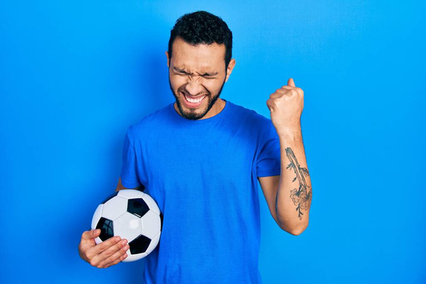 Latynos z brodą trzymający piłkę nożną bardzo szczęśliwy i podekscytowany wykonując gest zwycięzcy z podniesionymi ramionami, uśmiechnięty i wołający o sukces. koncepcja uroczystości.  - Zdjęcie, obraz