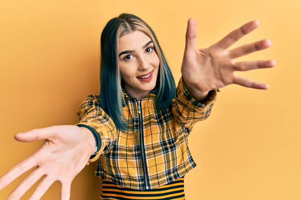 Young modern girl wearing casual yellow shirt looking at the camera smiling with open arms for hug. cheerful expression embracing happiness.  - Фото, изображение