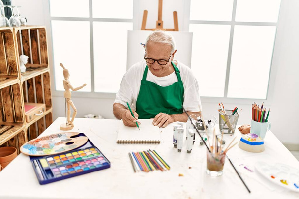 Senior grijsharige kunstenaar glimlachend gelukkig schilderij zittend op de tafel in kunststudio. - Foto, afbeelding