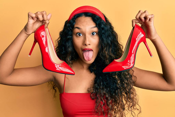 Young latin woman holding red high heel shoes sticking tongue out happy with funny expression.  - Photo, Image