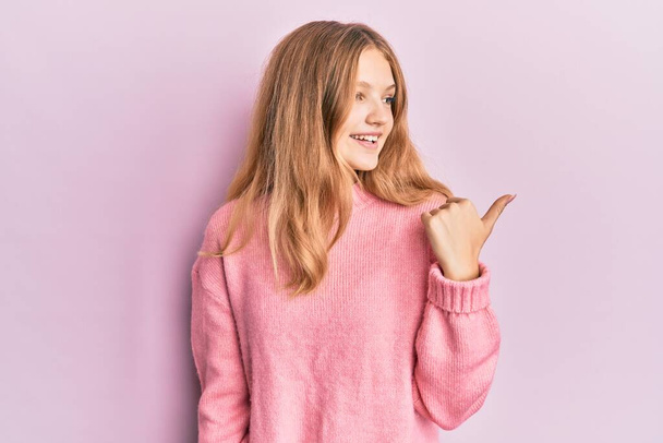 Beautiful young caucasian girl wearing casual clothes smiling with happy face looking and pointing to the side with thumb up.  - Fotografie, Obrázek