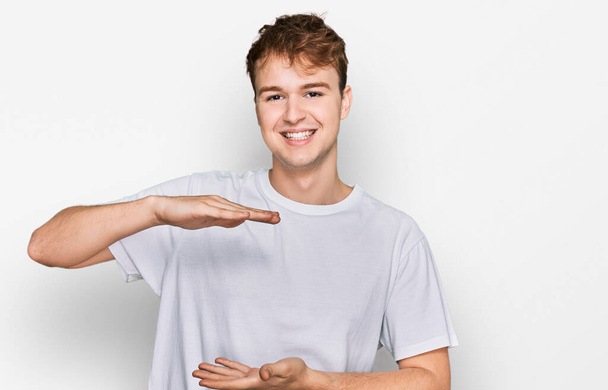 Jonge blanke man draagt casual wit t-shirt gebaren met handen tonen grote en grote maat teken, maat symbool. glimlachend kijkend naar de camera. meetconcept.  - Foto, afbeelding