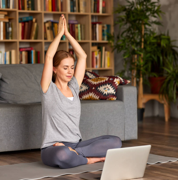 Young peaceful female in sportswear meditating with closed eyes, doing breath exercises while having yoga class online on laptop at home, full length. Mindful woman enjoying activity during quarantine - Foto, immagini
