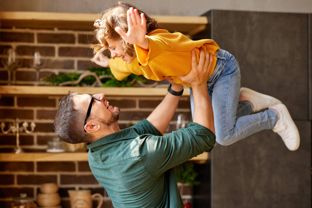 Playtime with dad. Little excited girl daughter flying in air in fathers arms while spending time together and having fun at home, young happy family daddy and child playing together indoors - Photo, Image