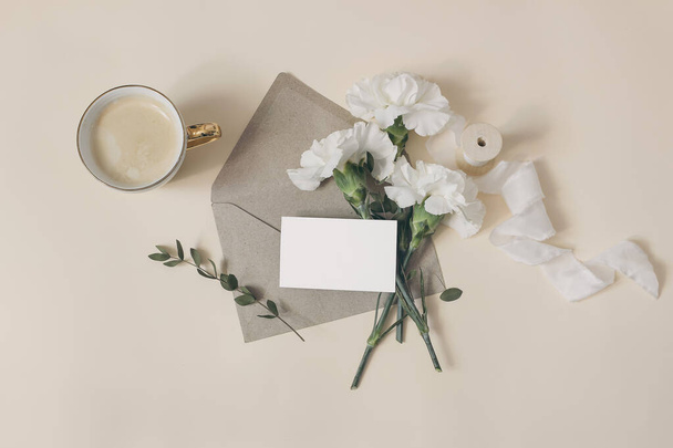 Feminine breakfast composition. Business card mockup. Wedding or birthday stationery. Cup of coffee, white carnation flowers and eucalyptus branches. Beige table background. Flat lay, top view. - 写真・画像