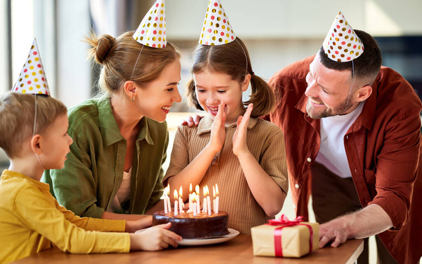 Heyecanlı küçük kız çikolatalı pastaya mum üflerken ve doğum gününü evinde mutlu bir aileyle kutlarken, parti şapkalı pozitif ebeveynler kızını tebrik ederken - Fotoğraf, Görsel