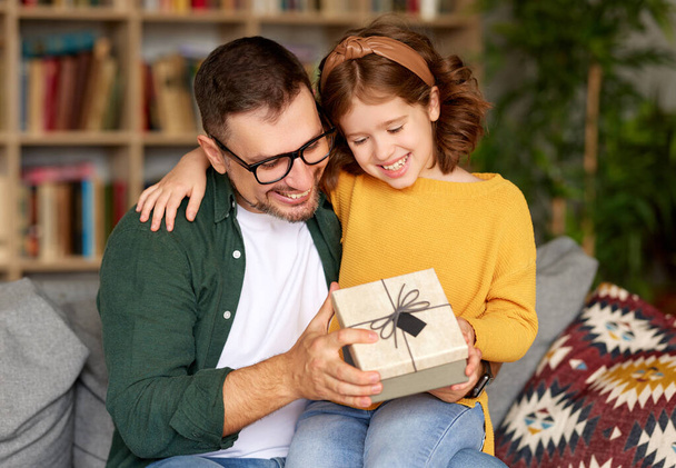 Christmas surprise for beloved dad. Cute little girl daughter receiving wrapped gift box from daddy on holidays while sitting on fathers knees, looking at him and smiling. Family celebration concept - Foto, Bild