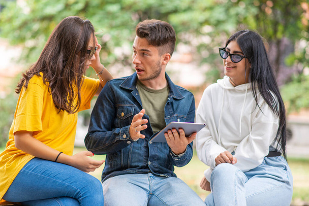 Three students studying together with a digital tablet sitting on a bench outdoor - Foto, imagen