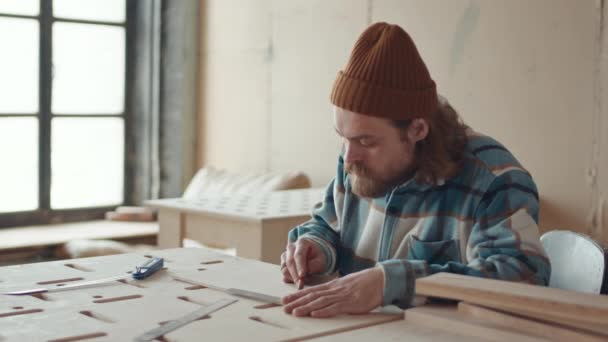 Orta boy erkek sakallı beyaz marangoz günlük kıyafetler giyer marangozluk atölyesinde tezgahta oturur ve kalem kullanarak ahşap bankta resim çizer. - Video, Çekim