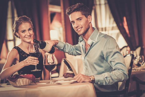 Beautiful young couple with glasses of red wine in luxury restaurant  - Фото, изображение