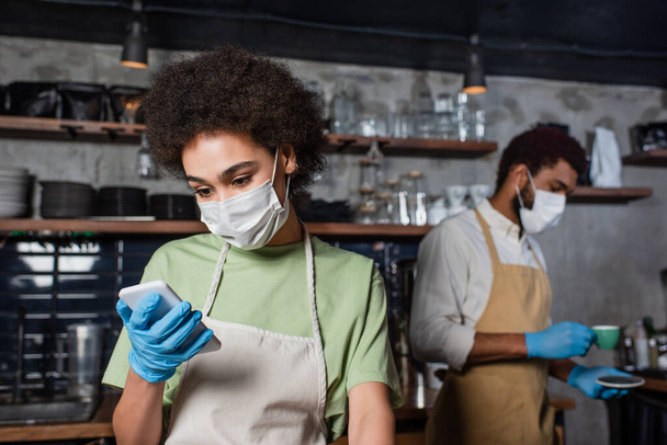 Afrikaanse amerikaanse barista in medisch masker en latex handschoen met behulp van smartphone in de buurt van collega in cafe  - Foto, afbeelding