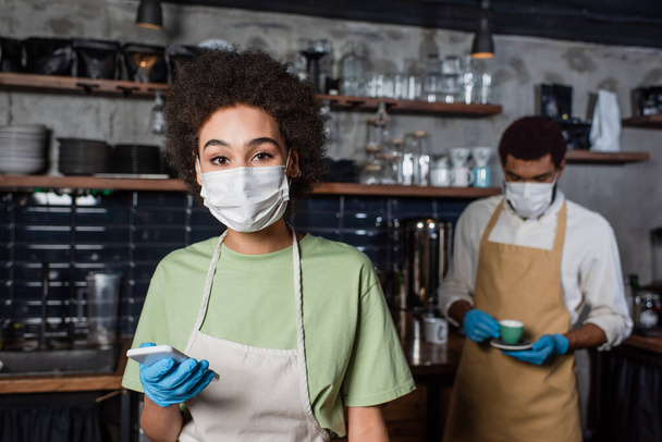 African american barista in medical mask and latex glove holding cellphone near blurred colleague in cafe  - Photo, Image