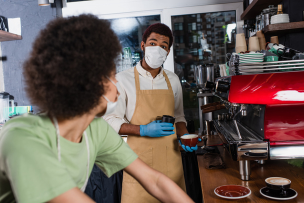 Barista afroamericano en máscara médica y guantes de látex haciendo café cerca de colega borroso en la cafetería  - Foto, imagen