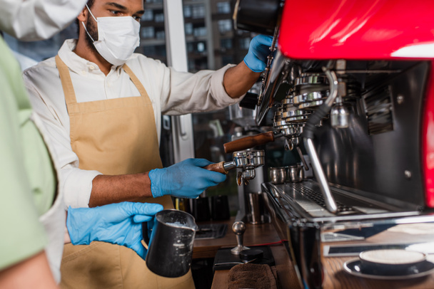 Tıbbi maskeli Afrikalı Amerikalı barista kahve makinesinde bulanık meslektaşının yanında kahve yapıyor.  - Fotoğraf, Görsel