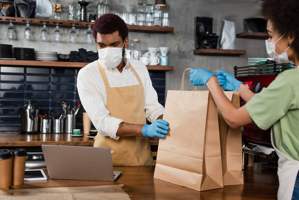 Afrikaans-Amerikaanse barista in medisch masker geven papieren zakken aan collega in de buurt laptop in cafe  - Foto, afbeelding