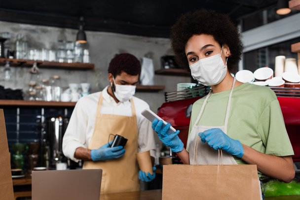 African american barista in medical mask and latex gloves using smartphone and holding paper bag near colleague  - Fotó, kép