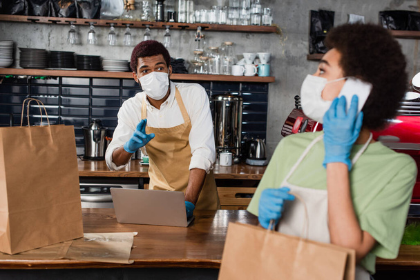 African american barista in medical mask pointing with fingers near colleague with paper bag talking on smartphone  - Φωτογραφία, εικόνα