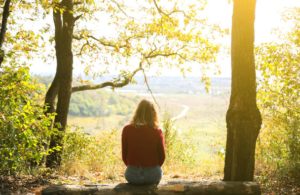 Stílusos hipszter lány ül az erdőben. Boldog nő élvezi az őszi természetet. Gyönyörű táj. Életmód utazási koncepció. Sötétvörös pulcsi. Sárga levelek a fákon. Kilátás a hegyről. - Fotó, kép