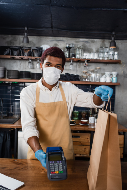 Afrikai amerikai barista orvosi maszkban és latex kesztyű kezében fizetési terminál és papírzacskó  - Fotó, kép