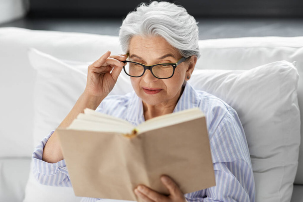 ηλικιωμένη γυναίκα με γυαλιά διαβάζοντας βιβλίο στο κρεβάτι στο σπίτι - Φωτογραφία, εικόνα